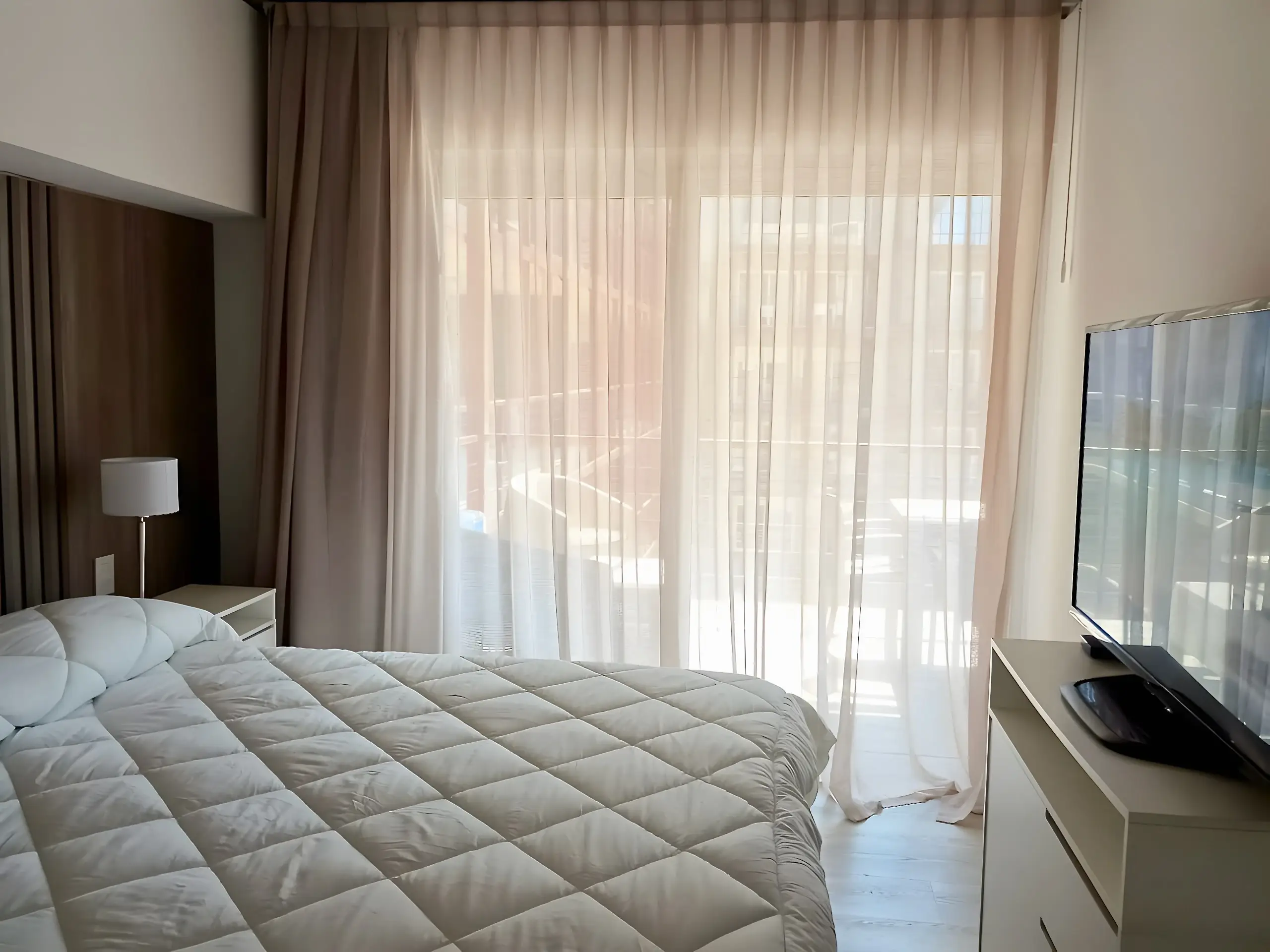 Dormitorio con smart TV, cortinas y ventanal al balcón